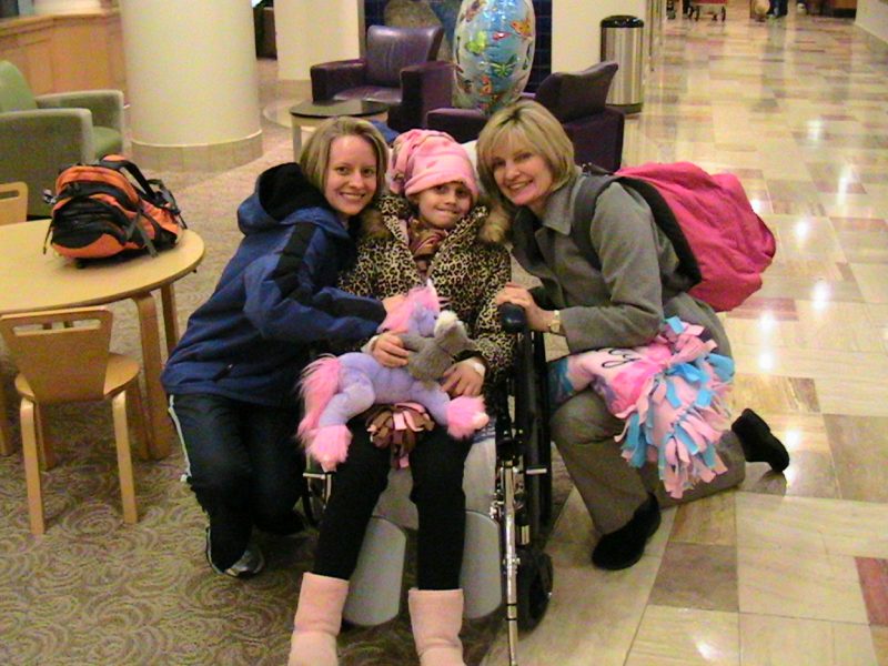 mobility | Batten Disease News | Laura and her mom, Sharon, kneel next to Laura's sister, Taylor, who is seated in a wheelchair wearing a cheetah-print jacket, pink boots, and a pink hat. Taylor has just been discharged from the hospital following a neural stem cell transplant.
