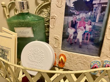 A close-up photo shows a small metal shelf hanging in a bathroom. It holds several bath products as well as a framed family photo. The frame is designed to appear sandy, with footprints going up the sides and colorful flip-flops at the bottom. In the photo, three women – the author's mother, grandmother, and younger sister – sit on a weathered bench swing.
