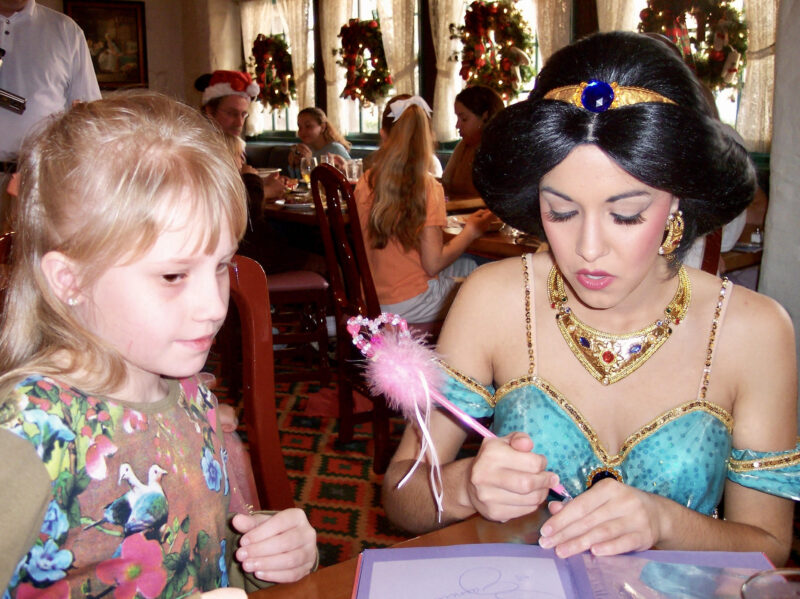 A young girl in a floral dress sits at a table next to a woman dressed as the Disney character Princess Jasmine, who holds a large pink pen and is signing an autograph for the girl. They are indoors at a themed restaurant at Disney World. 